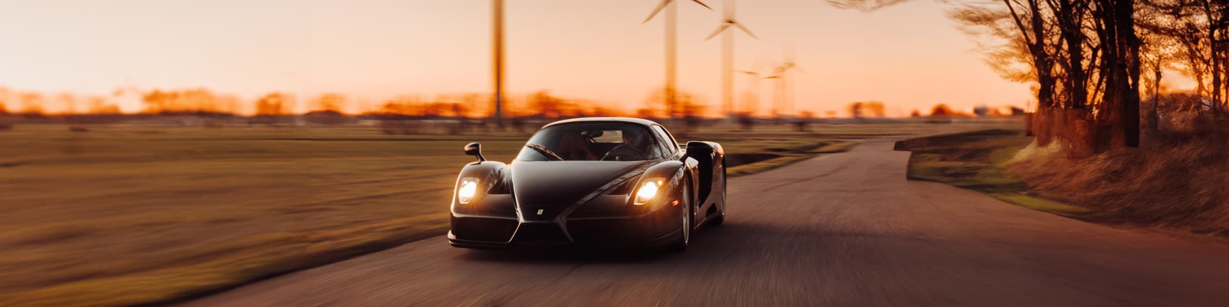 A black sports car drives down a rural road at sunset, with wind turbines and blurred trees in the background.